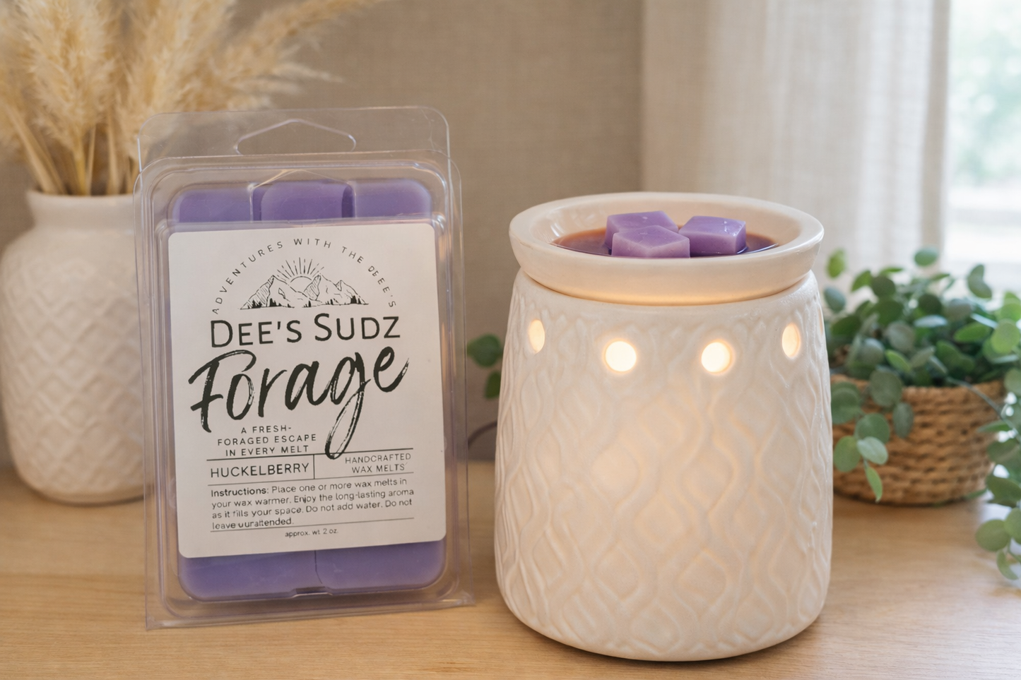 Clear plastic clamshell wax melt package with Dee’s Sudz Forage Huckleberry label beside a white ceramic electric wax melt lamp, featuring purple wax melt cubes warming in the dish under soft bulb light in a neutral boho setting with greenery and pampas grass.