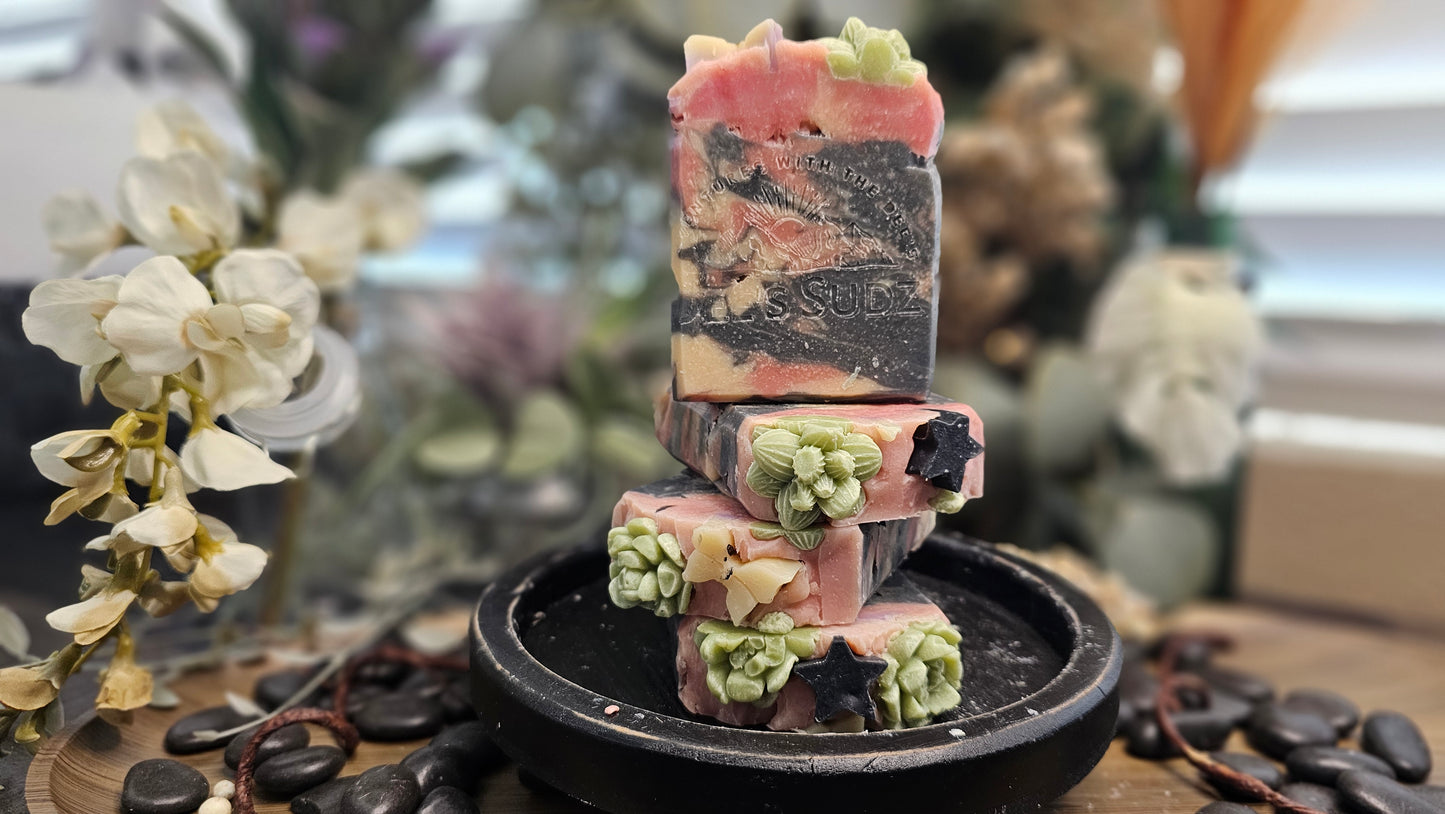 Rectangular handmade soap bars with the Dee’s Sudz logo, featuring a pink, black, and cream swirl design with decorative green, tan and black accents, stacked on a black stand with a natural blurred background.