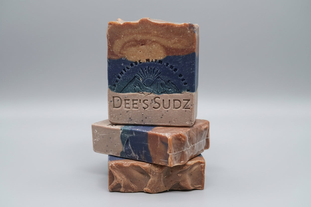 Rectangular handmade soap bar with the Dee’s Sudz logo, featuring layered tan, blue, and orange tones with a subtle swirl pattern, stacked on matching soap pieces with a grey background.