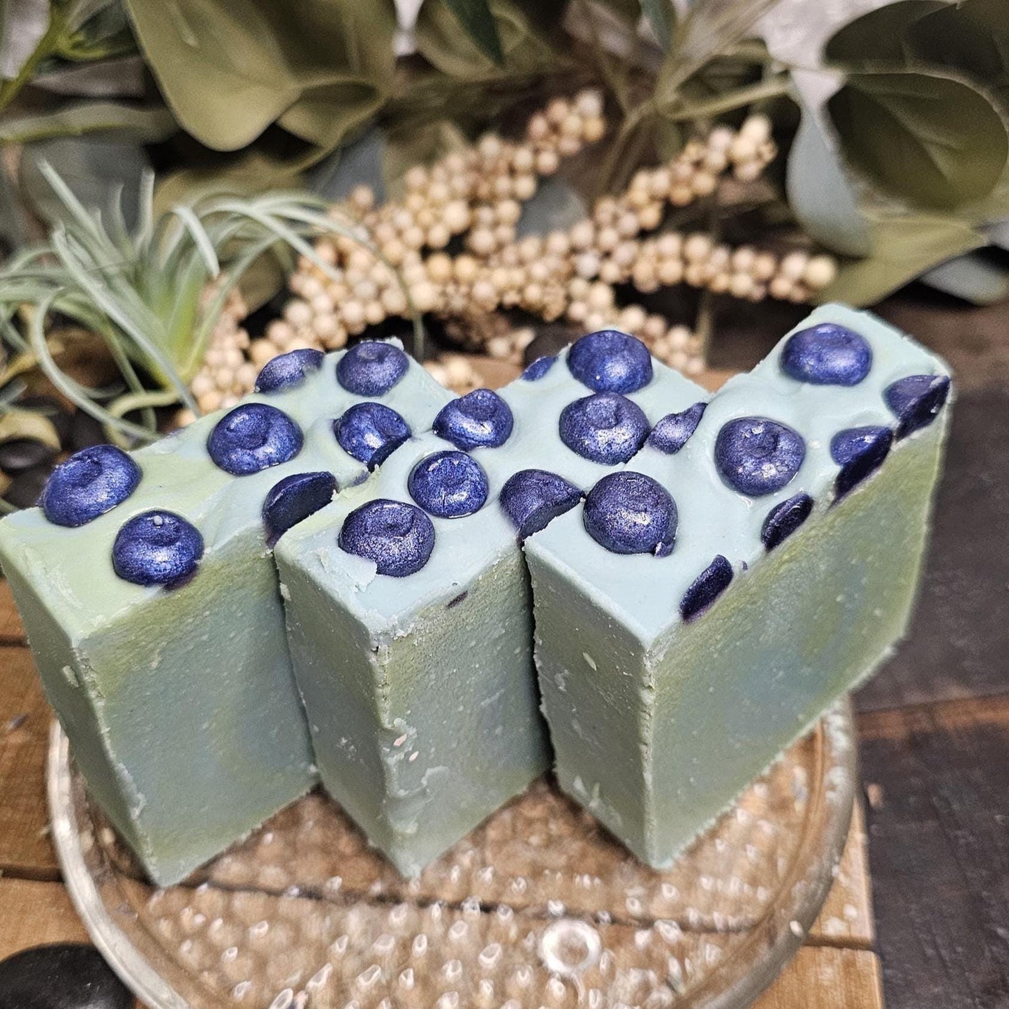 Rectangular handmade soap bars, featuring a soft green color with raised dark blue circular blueberry accents on top, arranged side by side on a glass dish.