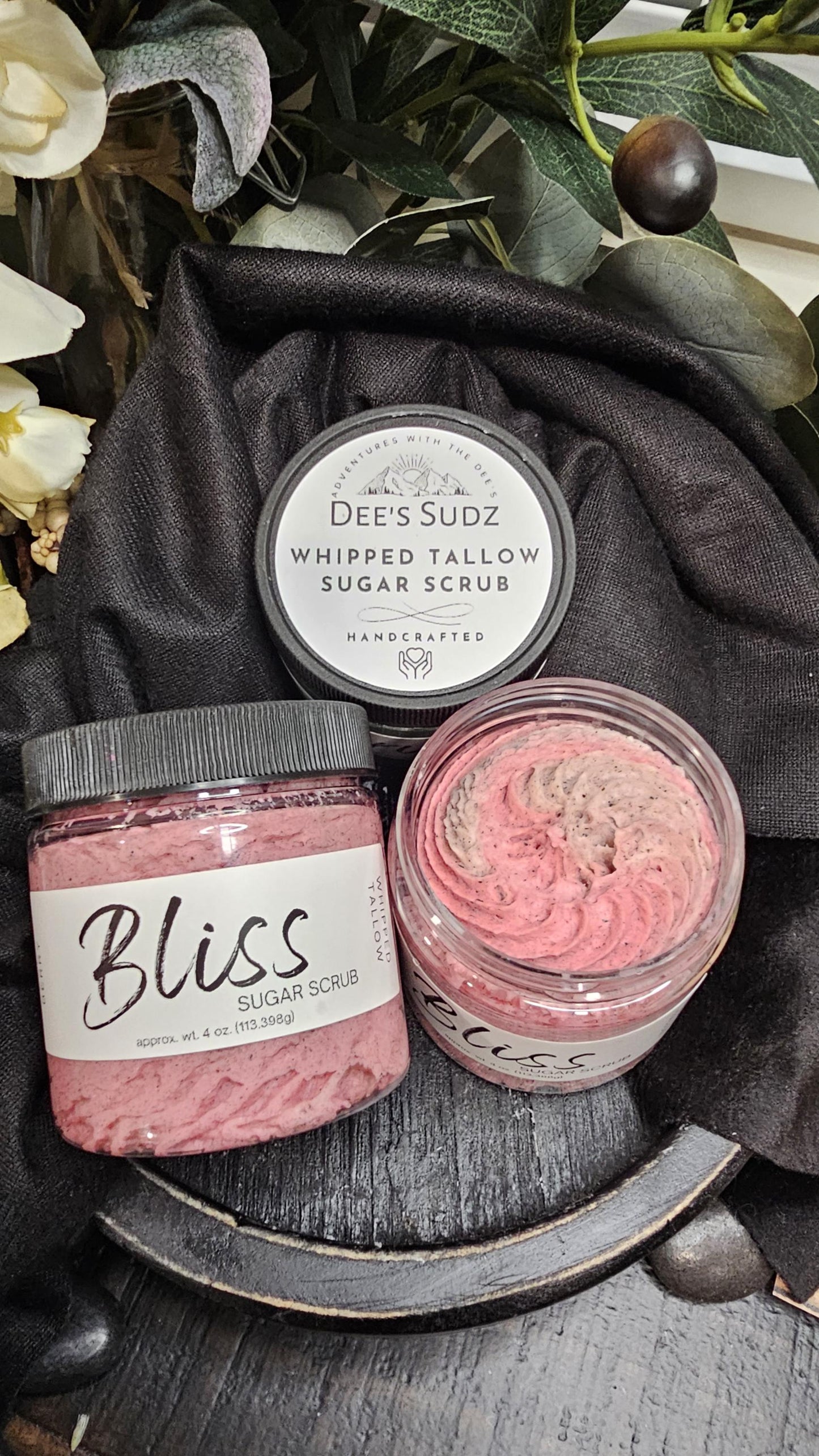 Three clear plastic jars of Dee’s Sudz whipped tallow sugar scrub with black lids, featuring a pink whipped texture and labeled containers displayed on a black wooden surface and a black cloth.