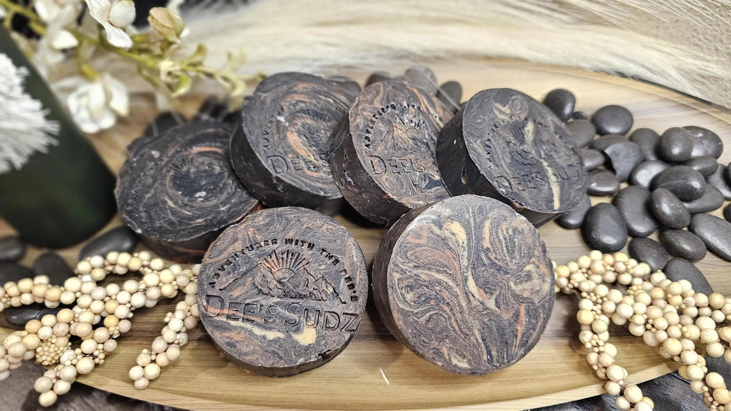 Round handmade soap bars with the Dee’s Sudz logo, featuring a charcoal base with tan, orange and cream swirl accents, arranged together on a wooden tray.