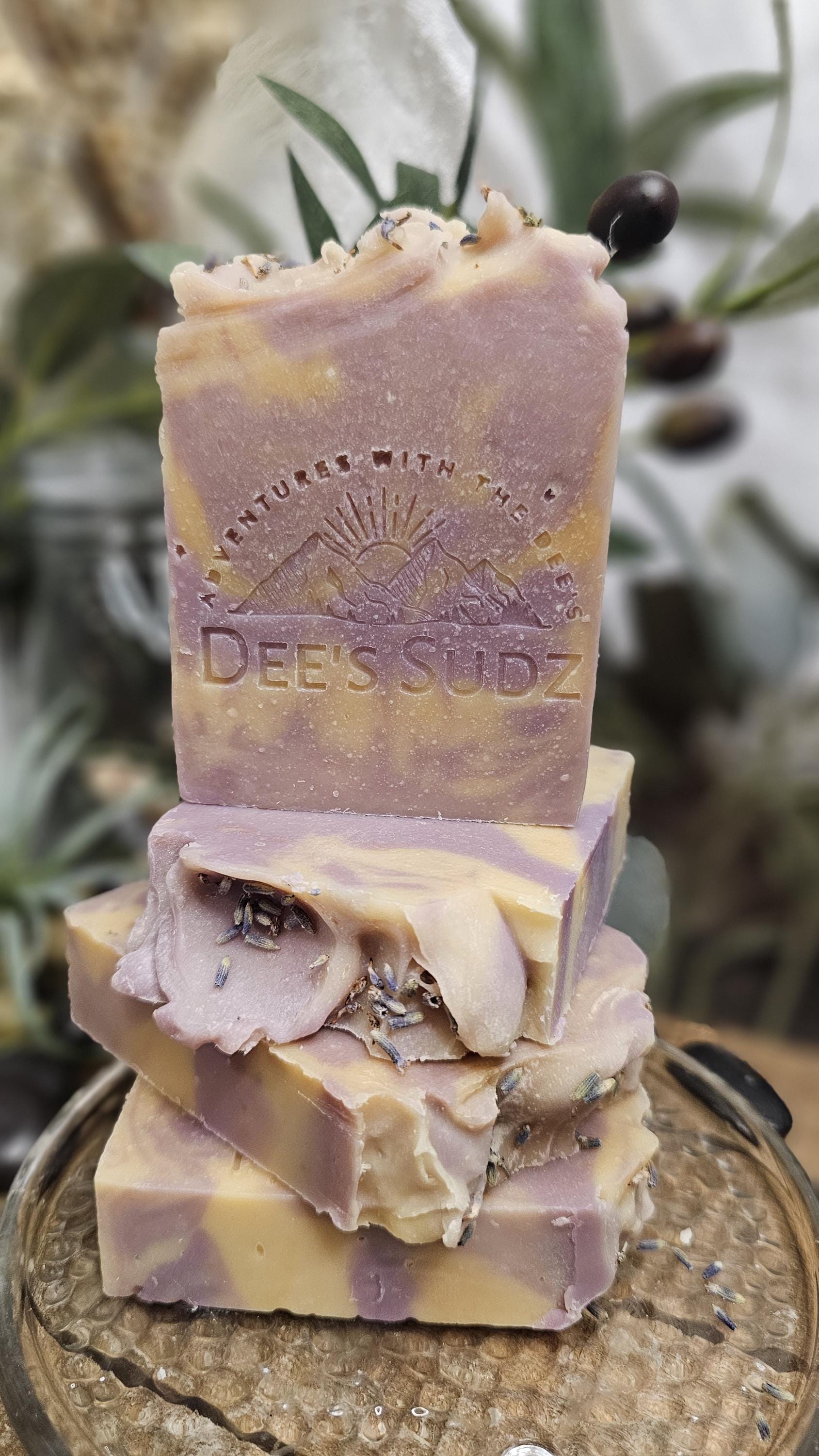 Rectangular handmade soap bar with the Dee’s Sudz logo, featuring a soft purple and cream swirl design with light botanical flecks, stacked on matching soap pieces with a natural blurred background. 