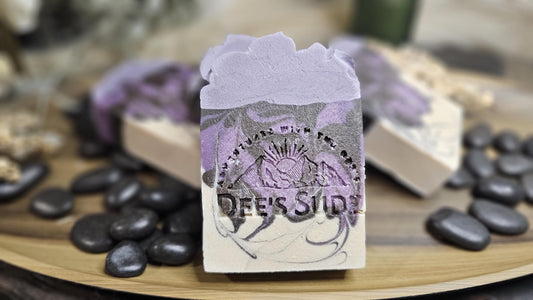 Rectangular handmade soap bar with the Dee’s Sudz logo, featuring a lavender, gray, and cream swirl design with a textured lavender top on a wooden surface with a blurred natural background.
