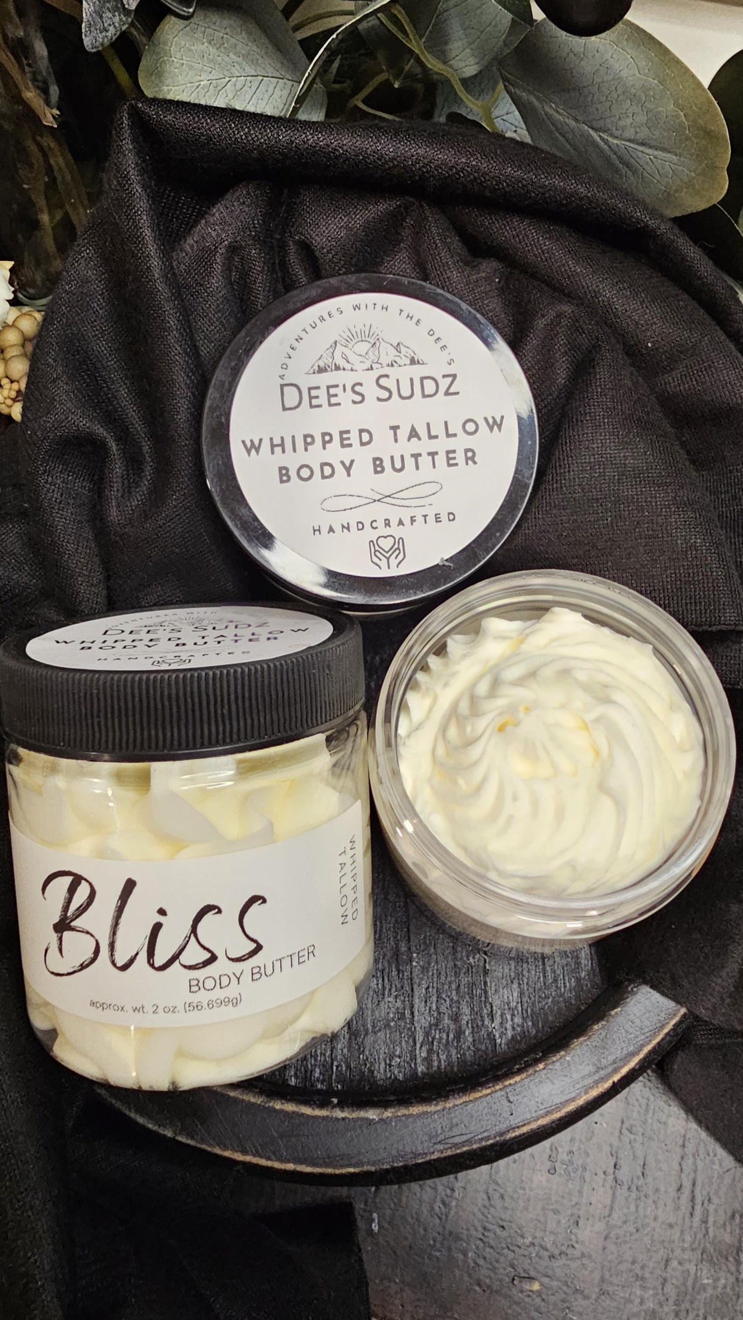 Three clear plastic jars of Dee’s Sudz whipped tallow body butter with black lids, featuring a white whipped texture and labeled BLISS displayed on a wooden surface.