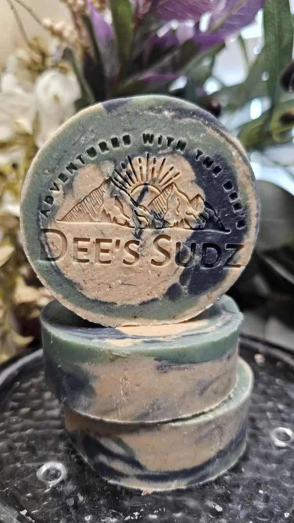 Round handmade soap bar with the Dee’s Sudz logo, featuring a natural green, blue and tan swirl design, stacked on matching soap bars.