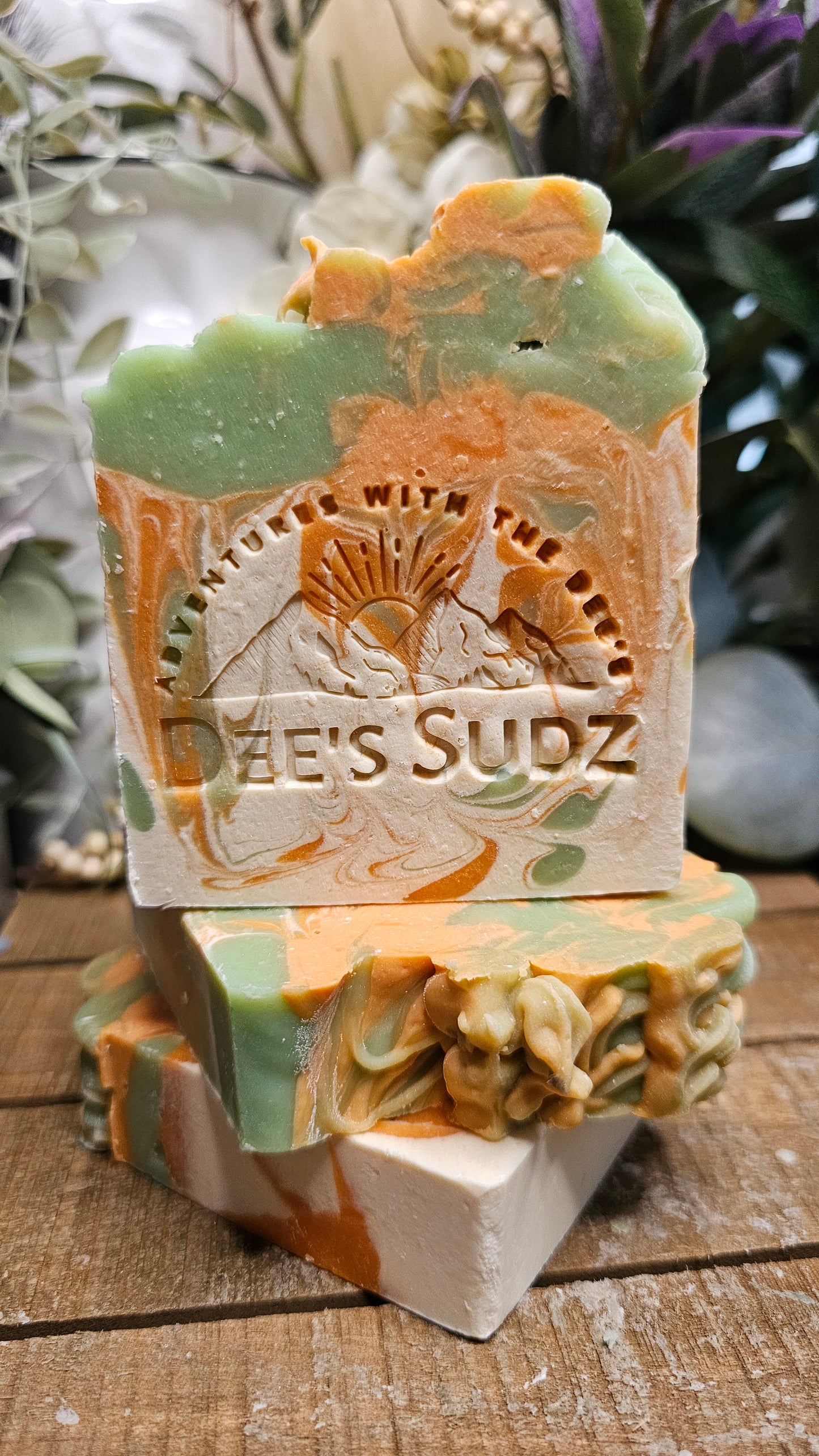 Rectangular handmade soap bar with the Dee’s Sudz logo, featuring a green, orange, and cream swirl design, stacked on matching soap pieces on a wood surface.