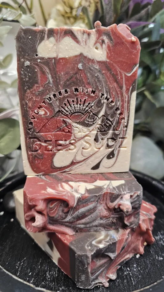 Rectangular handmade soap bar with the Dee’s Sudz logo, featuring a red, cream, and dark brown swirl design, stacked on matching soap pieces on a dark stand.