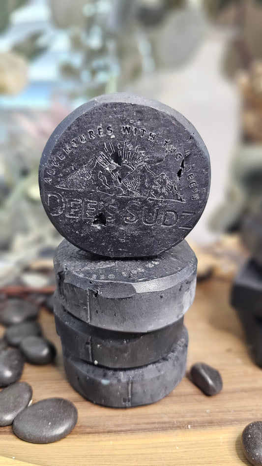 Round handmade soap bars with the Dee’s Sudz logo, featuring a solid charcoal black color, stacked vertically on a wood platform and a blurred natural background.