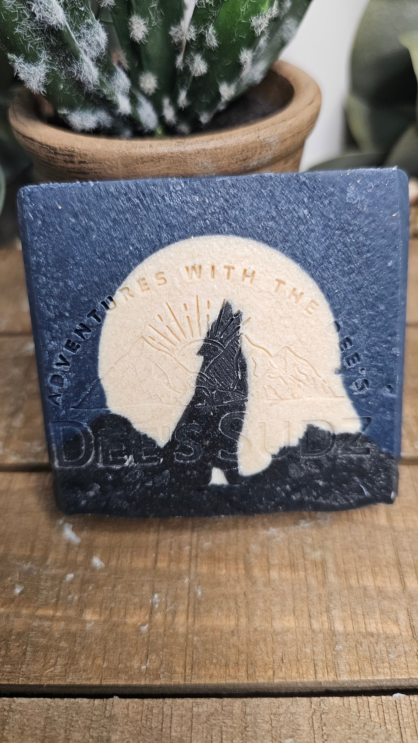 Rectangular handmade soap bar with the Dee’s Sudz logo, featuring a deep blue and cream color block design with a black wolf silhouette and moon graphic on the front displayed on a wood surface.