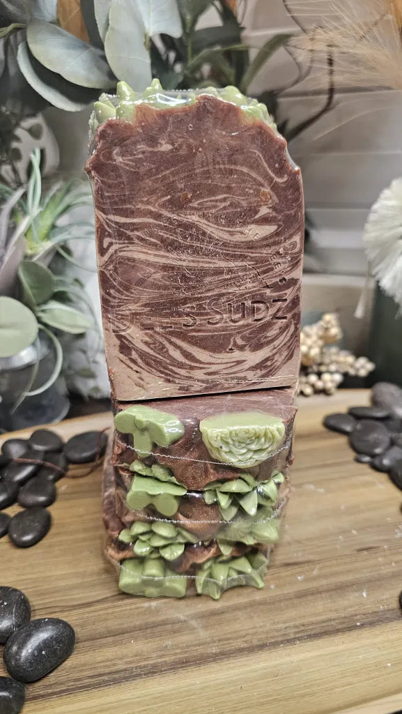 Rectangular handmade soap bar with the Dee’s Sudz logo, featuring a brown and cream swirl design with textured green decorative accents, stacked on matching soap pieces on a wood surface with decorative natural elements in the background.