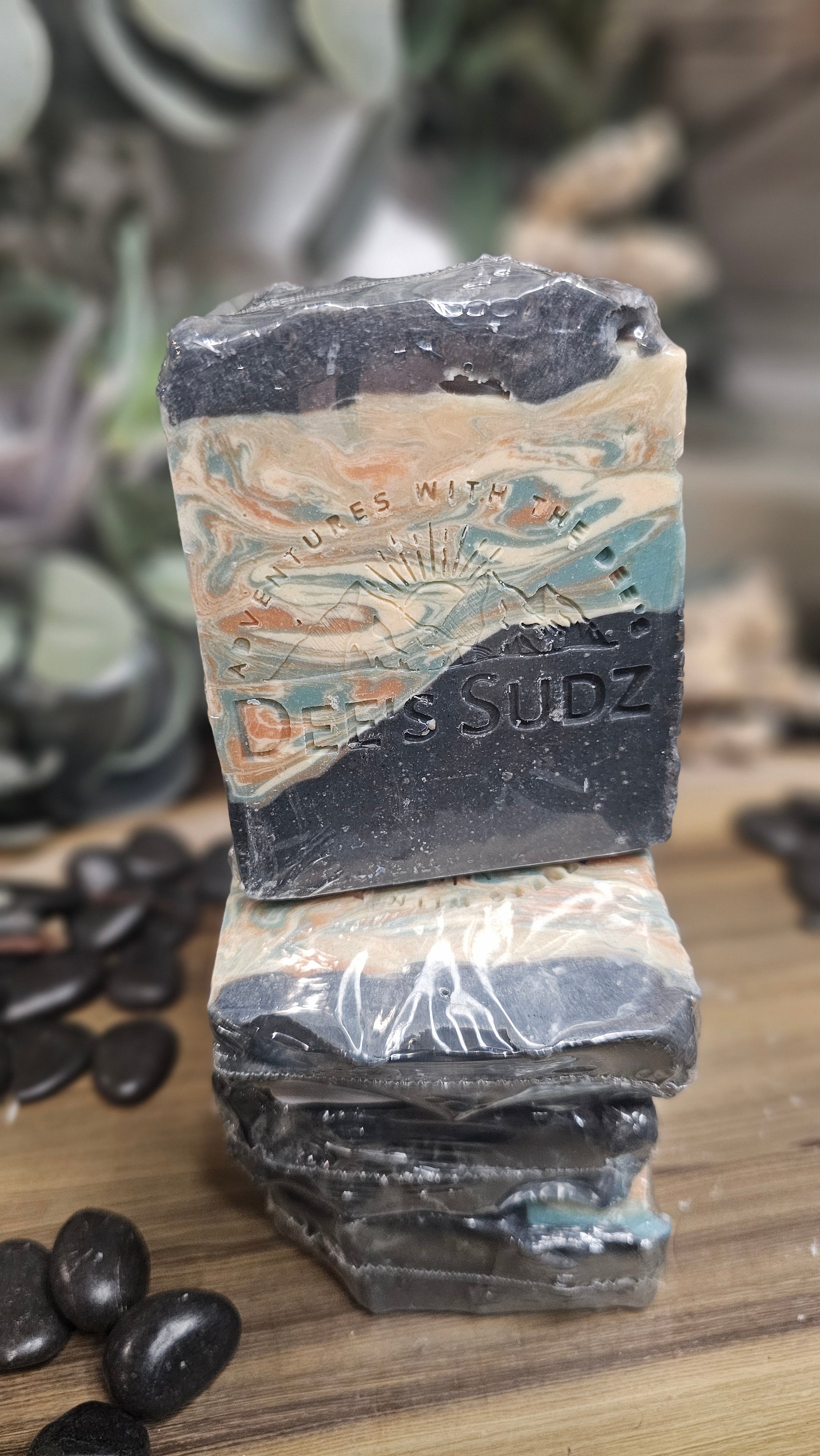 Rectangular handmade soap bar with the Dee’s Sudz logo, featuring a charcoal gray base with blue, tan, and cream swirl accents, stacked on matching soap pieces  on a wood surface with surrounding lack rocks. 