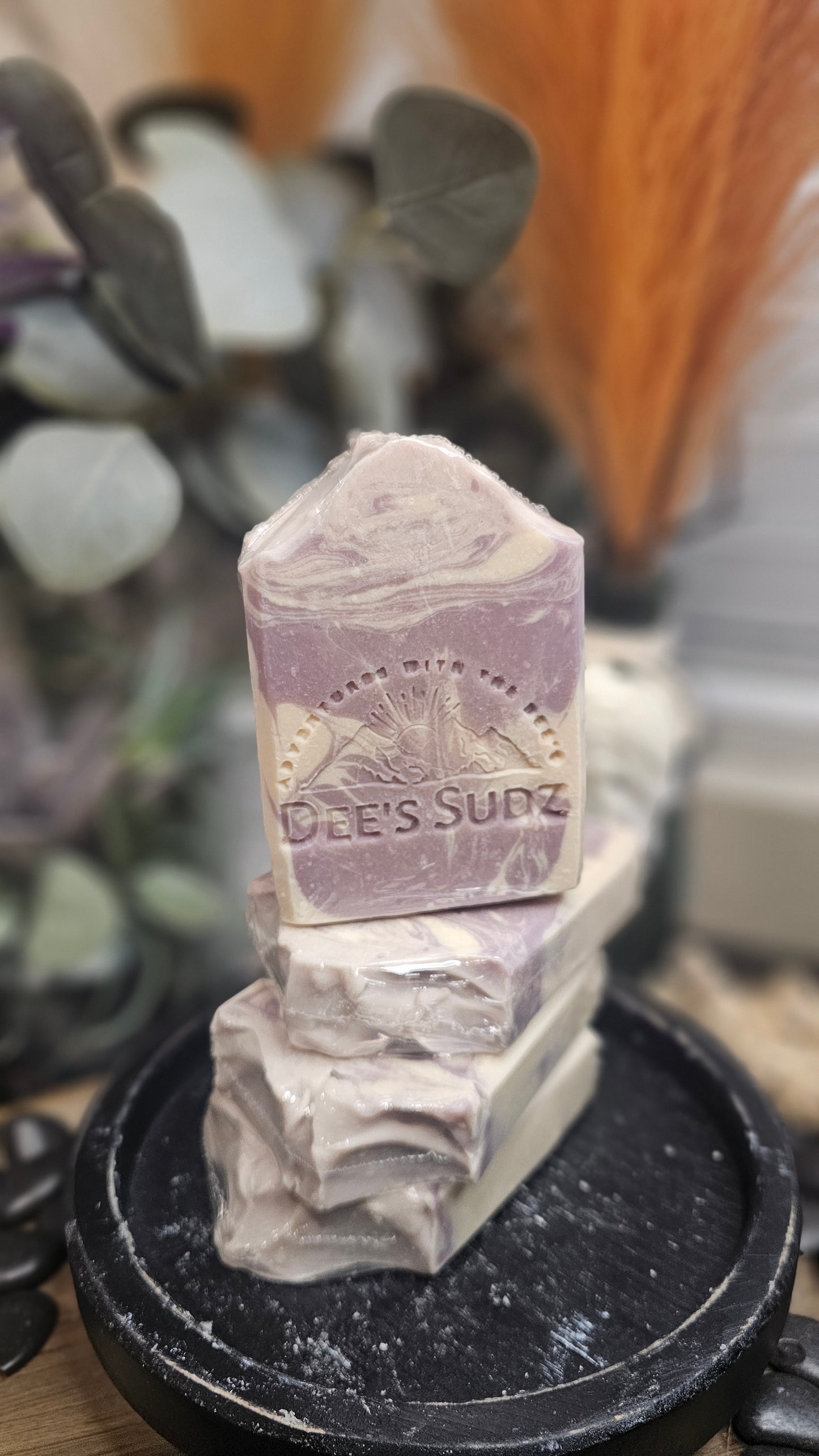 Rectangular handmade soap bar with the Dee’s Sudz logo, featuring a soft lavender and cream swirl design, stacked on matching soap pieces on a black stand with a blurred natural background.