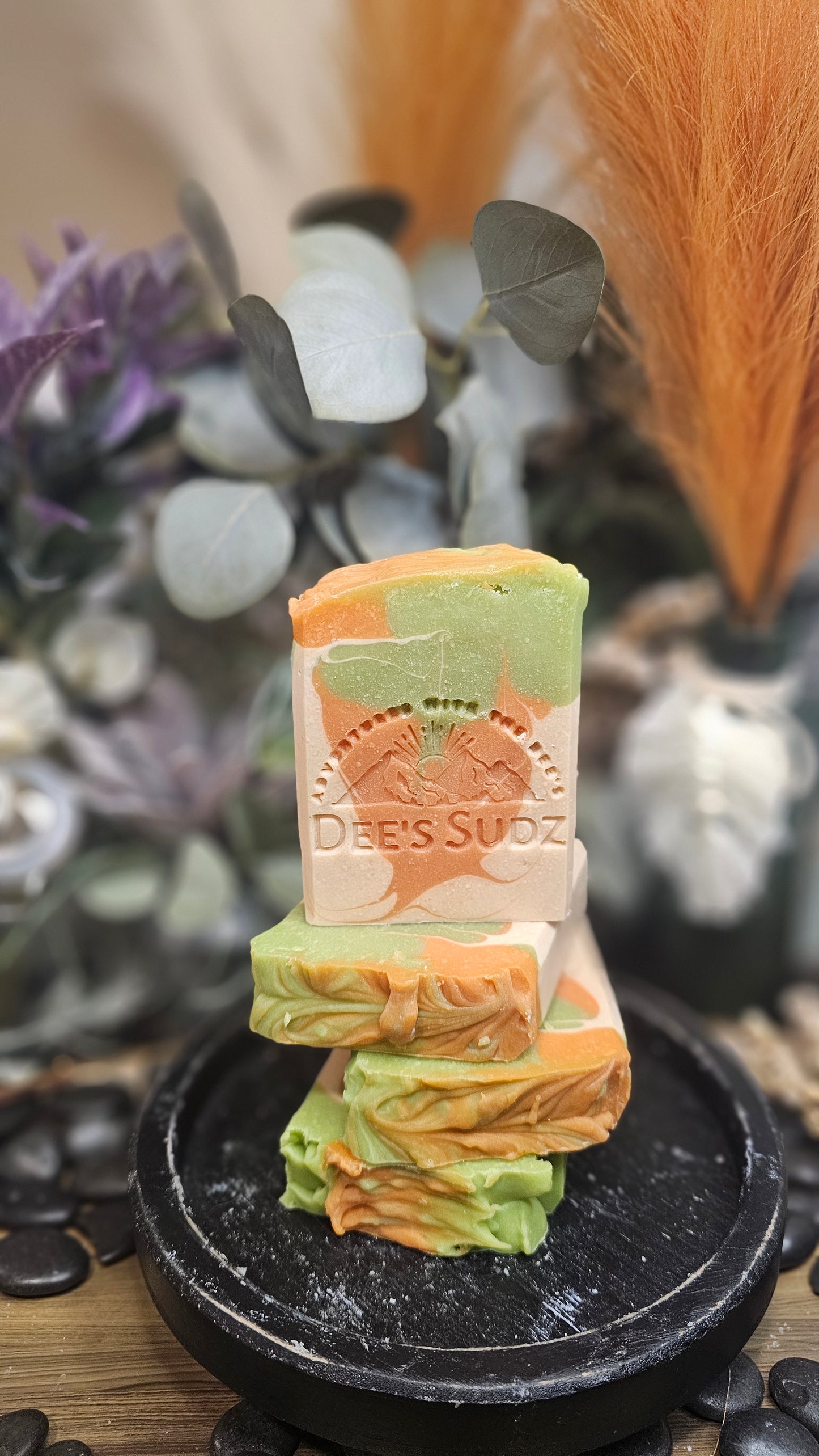 Rectangular handmade soap bar with the Dee’s Sudz logo, featuring a green, tan, and peach swirl design, stacked on matching soap pieces with a blurred natural background.