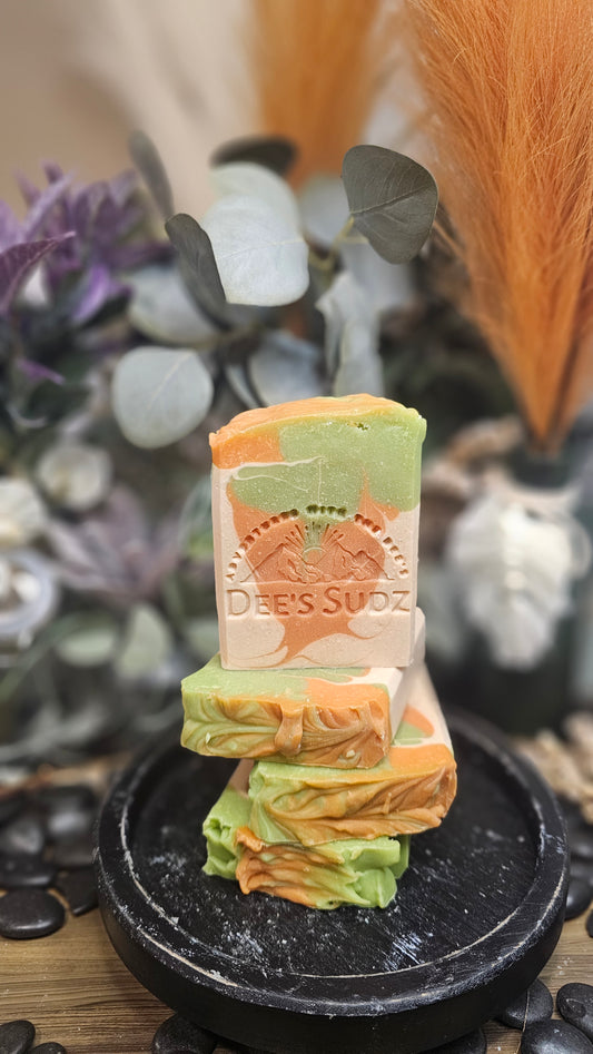 Rectangular handmade soap bar with the Dee’s Sudz logo, featuring a green, tan, and peach swirl design, stacked on matching soap pieces with a blurred natural background.