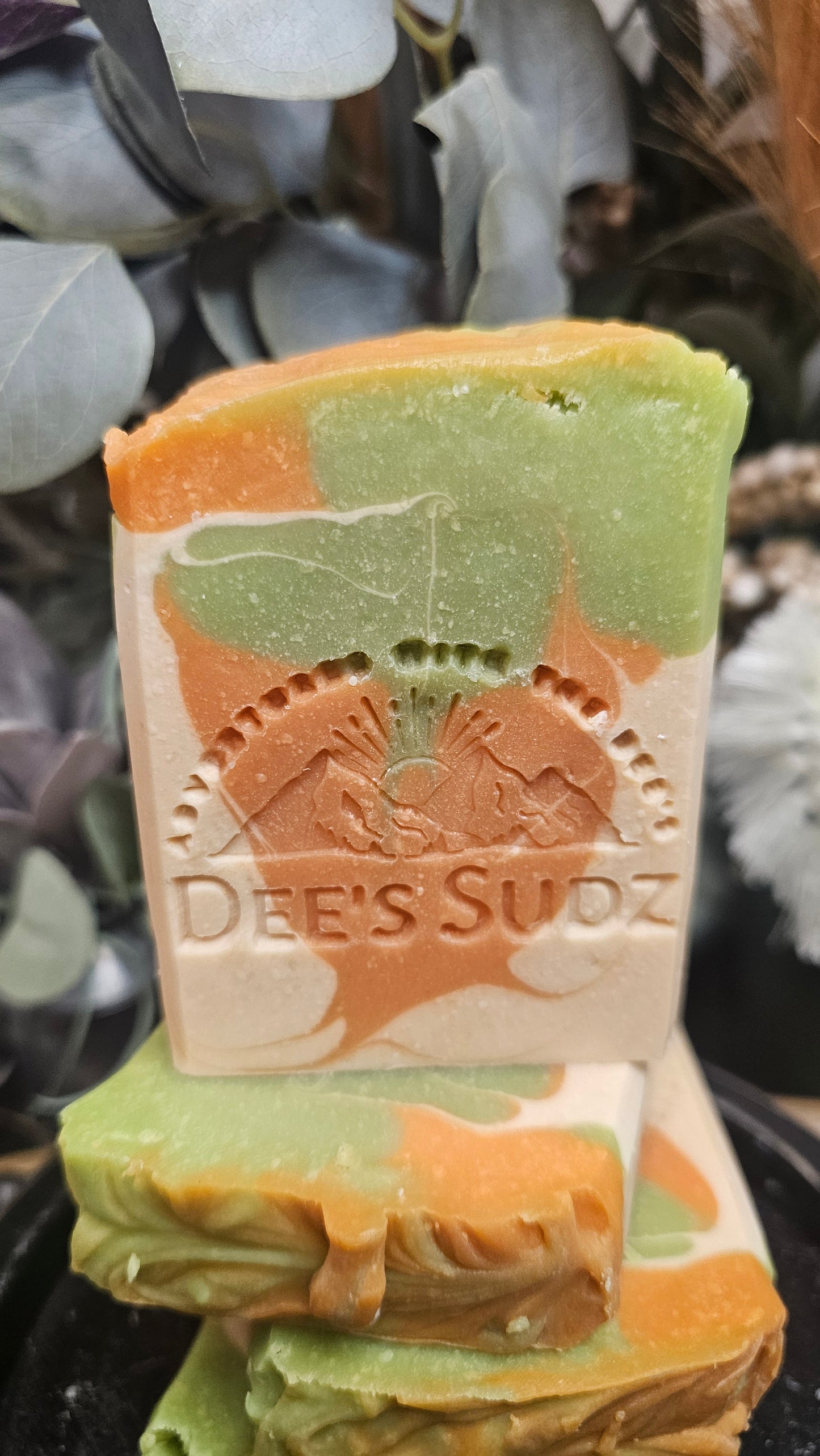Rectangular handmade soap bar with the Dee’s Sudz logo, featuring a green, tan, and peach swirl design, stacked on matching soap pieces with a blurred natural background.