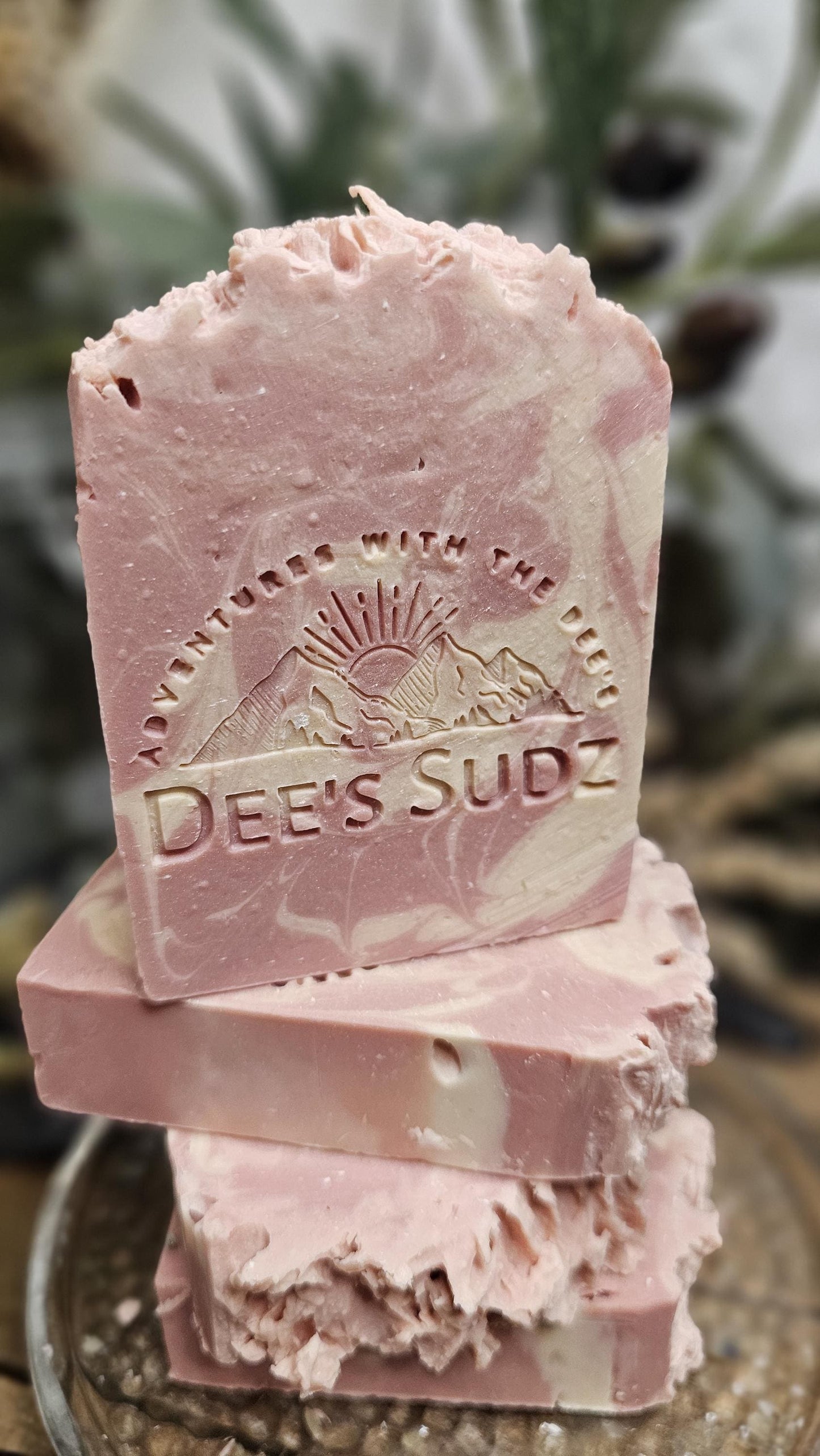 Rectangular handmade soap bar with the Dee’s Sudz logo, featuring soft blush pink and cream swirl tones with a textured top, stacked on matching soap pieces with a blurred natural background.