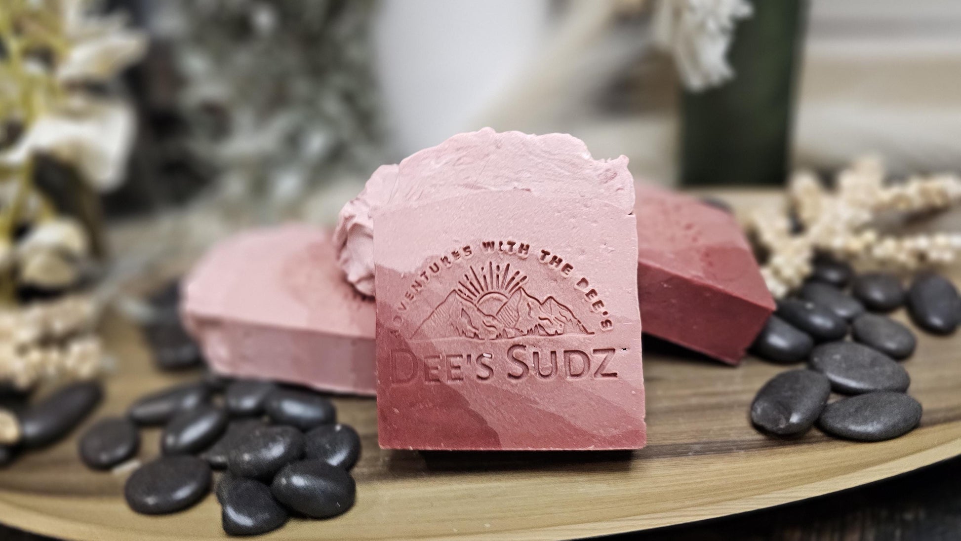 Rectangular handmade soap bar with the Dee’s Sudz logo, featuring layered blush pink and rose tones with a smooth, textured top on a wooden surface with stones and flowers.