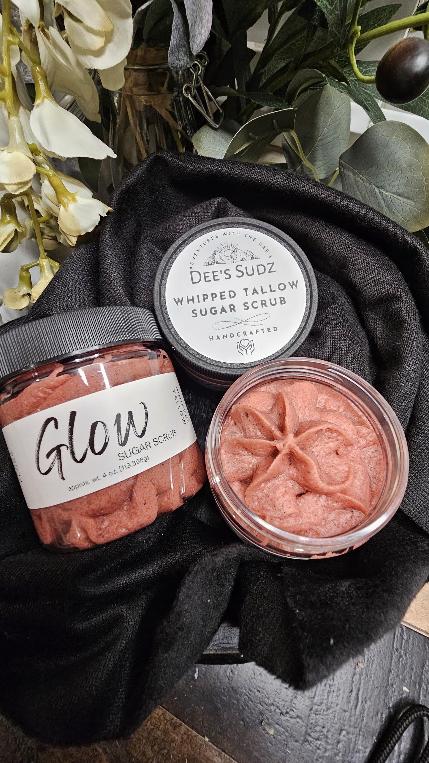 Three clear plastic jars of Dee’s Sudz whipped tallow sugar scrub with black lids, featuring a red whipped texture and labeled containers displayed on a black cloth.