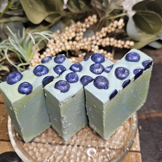 Rectangular handmade soap bars, featuring a soft green color with raised dark blue circular blueberry accents on top, arranged side by side on a glass dish.