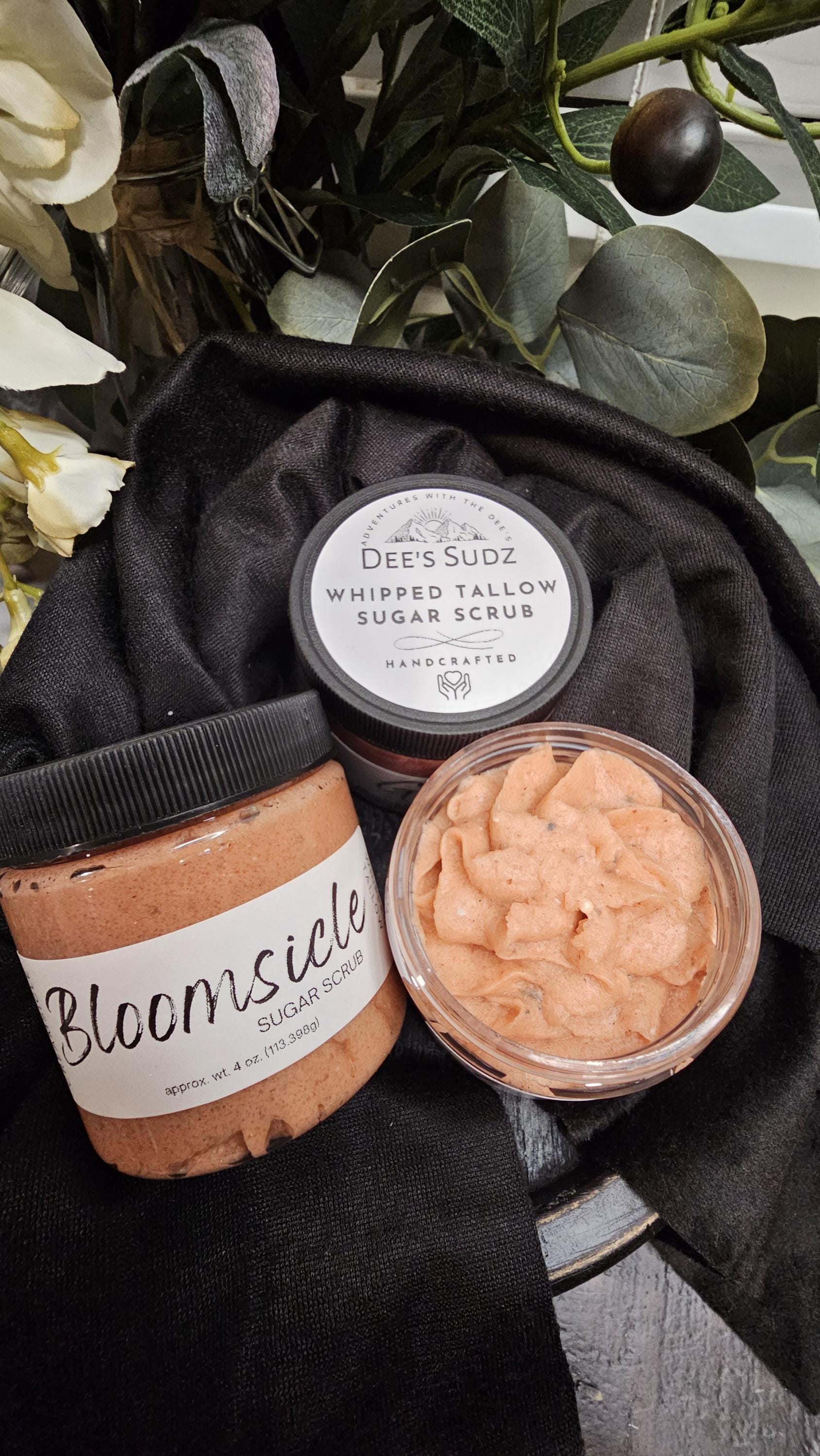 Three clear plastic jars of Dee’s Sudz whipped tallow sugar scrub with black lids, featuring a orange whipped texture and labeled containers displayed on a black cloth.