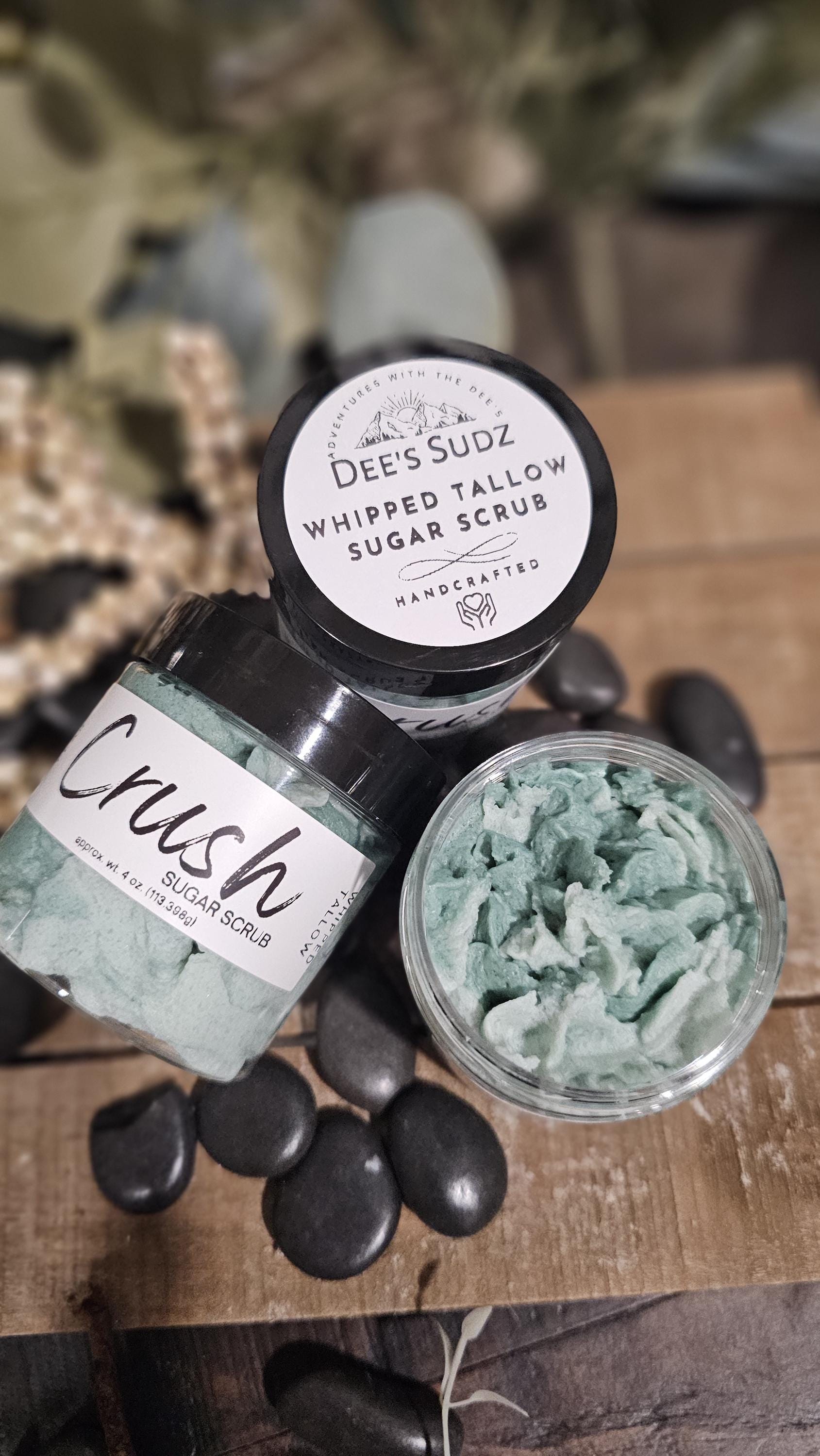 Three clear plastic jars of Dee’s Sudz whipped tallow sugar scrub with black lids, featuring a pale green whipped texture and labeled containers displayed on a wooden surface.