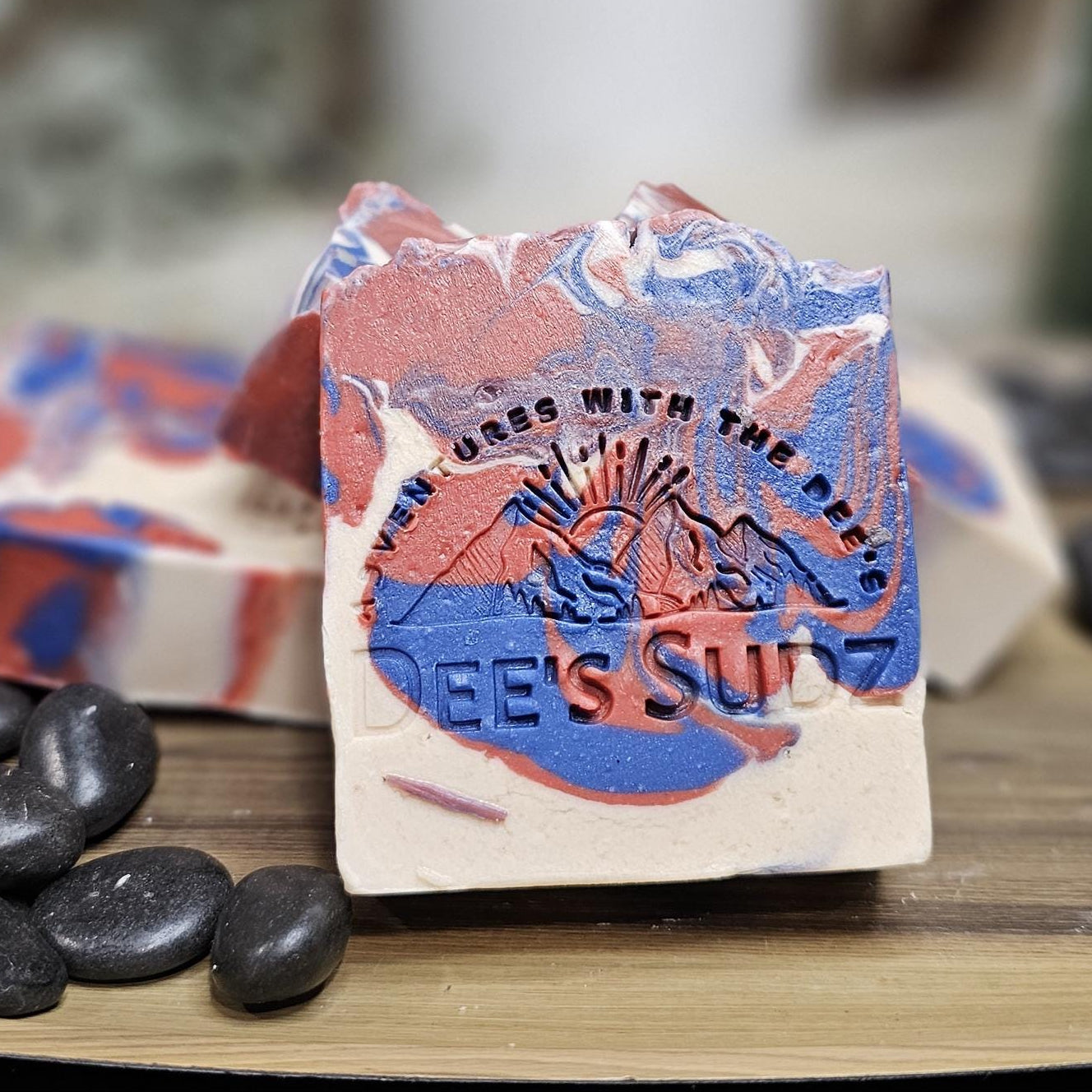 Rectangular handmade soap bar with the Dee’s Sudz logo, featuring a red, blue, and white swirl design with a textured top on a wood stand.
