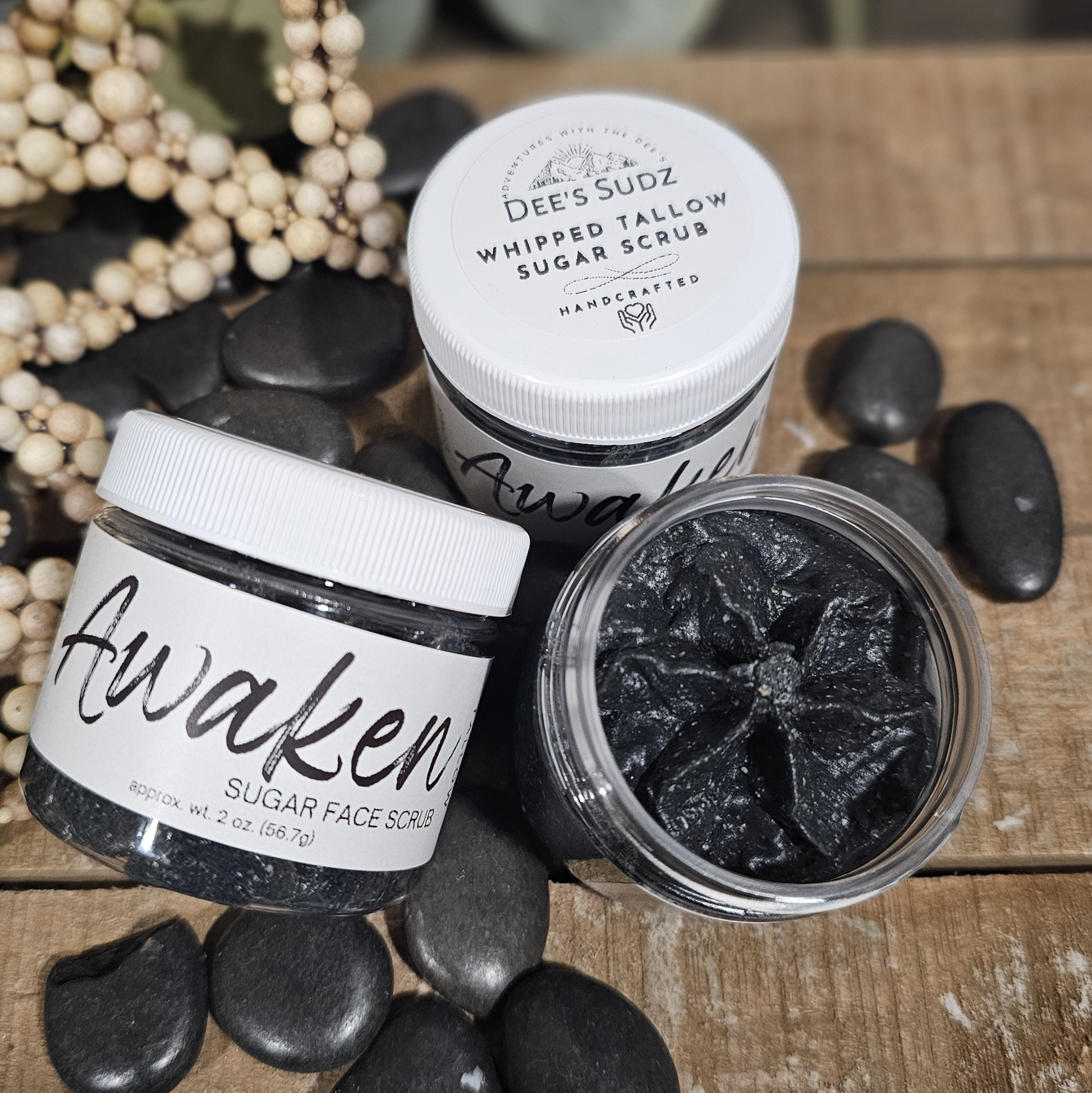 Open and closed jars of whipped tallow sugar face scrub by Dee’s Sudz, featuring the Awaken label, with a dark charcoal-colored scrub styled on a rustic wooden surface with black stones.