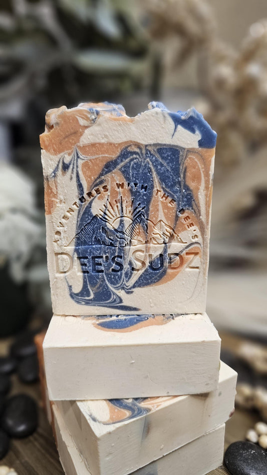 Rectangular handmade soap bar with the Dee’s Sudz logo, featuring a cream base with blue and orange swirl accents, stacked on matching soap pieces on a wooden surface