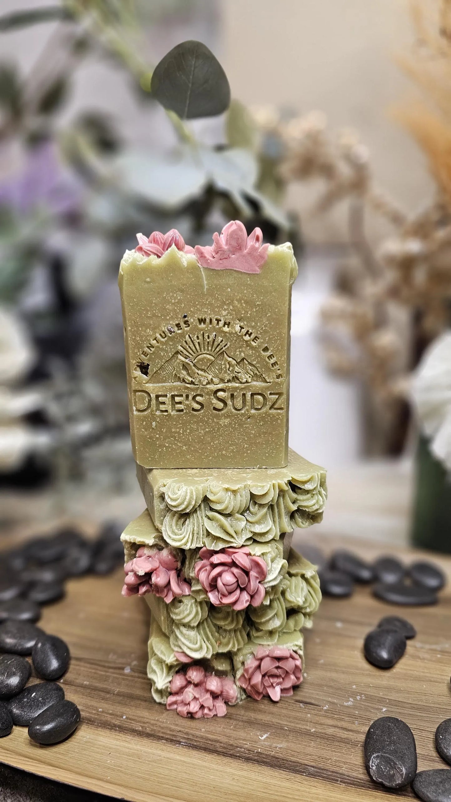 Rectangular handmade soap bar with the Dee’s Sudz logo, featuring a soft green base with pink floral decorative accents and a textured top, stacked on matching soap pieces on a wood surface.