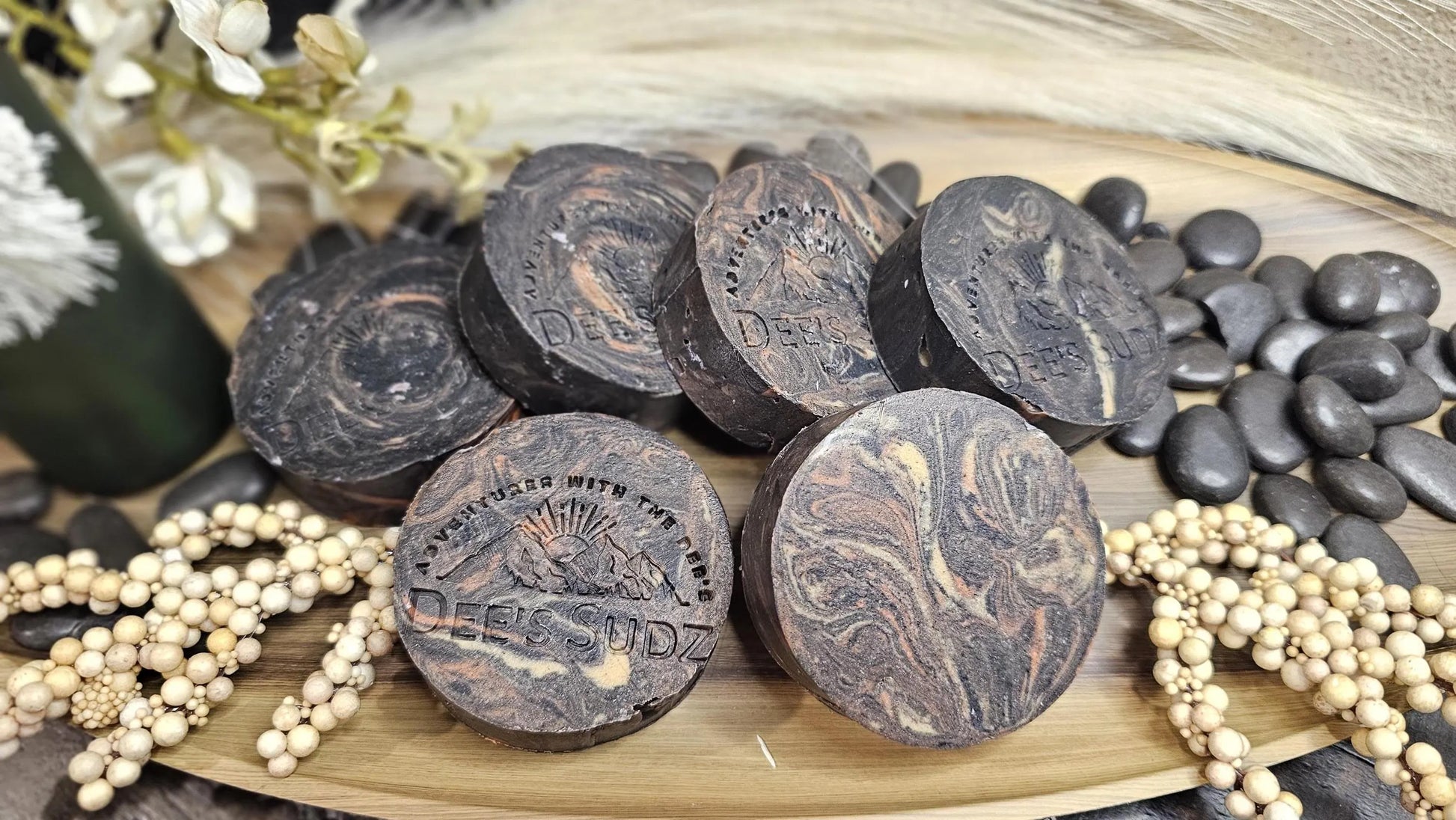 Round handmade soap bars with the Dee’s Sudz logo, featuring a charcoal base with tan, orange and cream swirl accents, arranged together on a wooden tray.