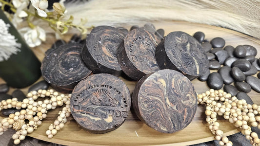 Round handmade soap bars with the Dee’s Sudz logo, featuring a charcoal base with tan, orange and cream swirl accents, arranged together on a wooden tray.